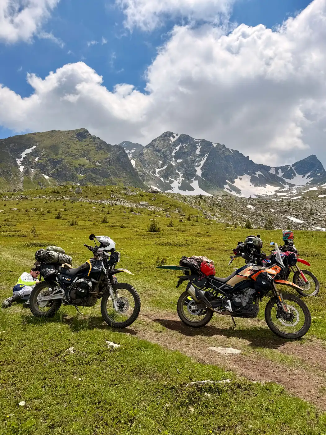 Two loaded adventure bikes on an alpine meadow below snow-patched limestone peaks