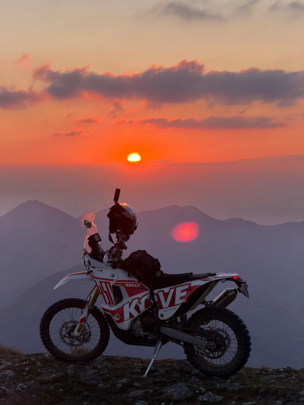 Kove 450 Rally on a ridge at sunset, deep orange sky over layered peaks