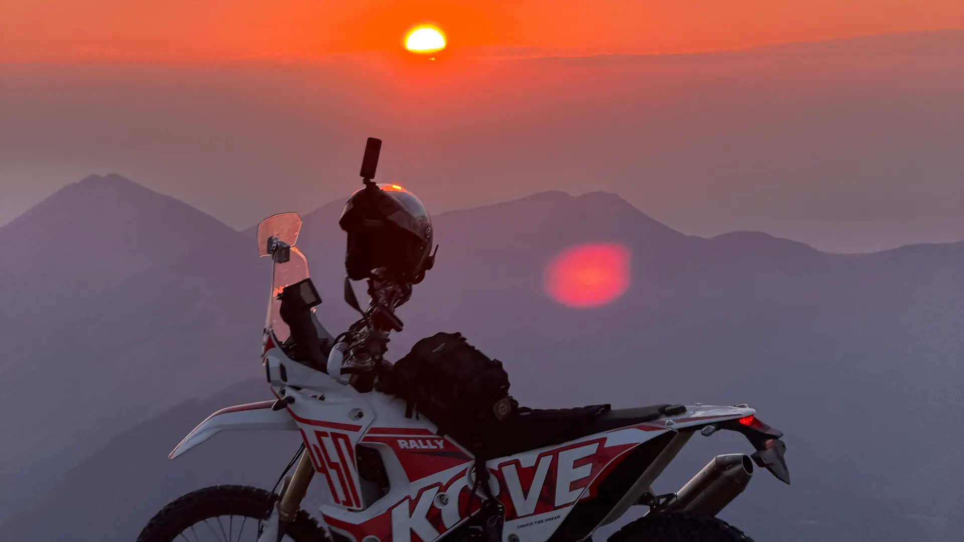 A Kove 450 Rally parked on a mountain summit at sunset, layered ridgelines fading into haze behind it