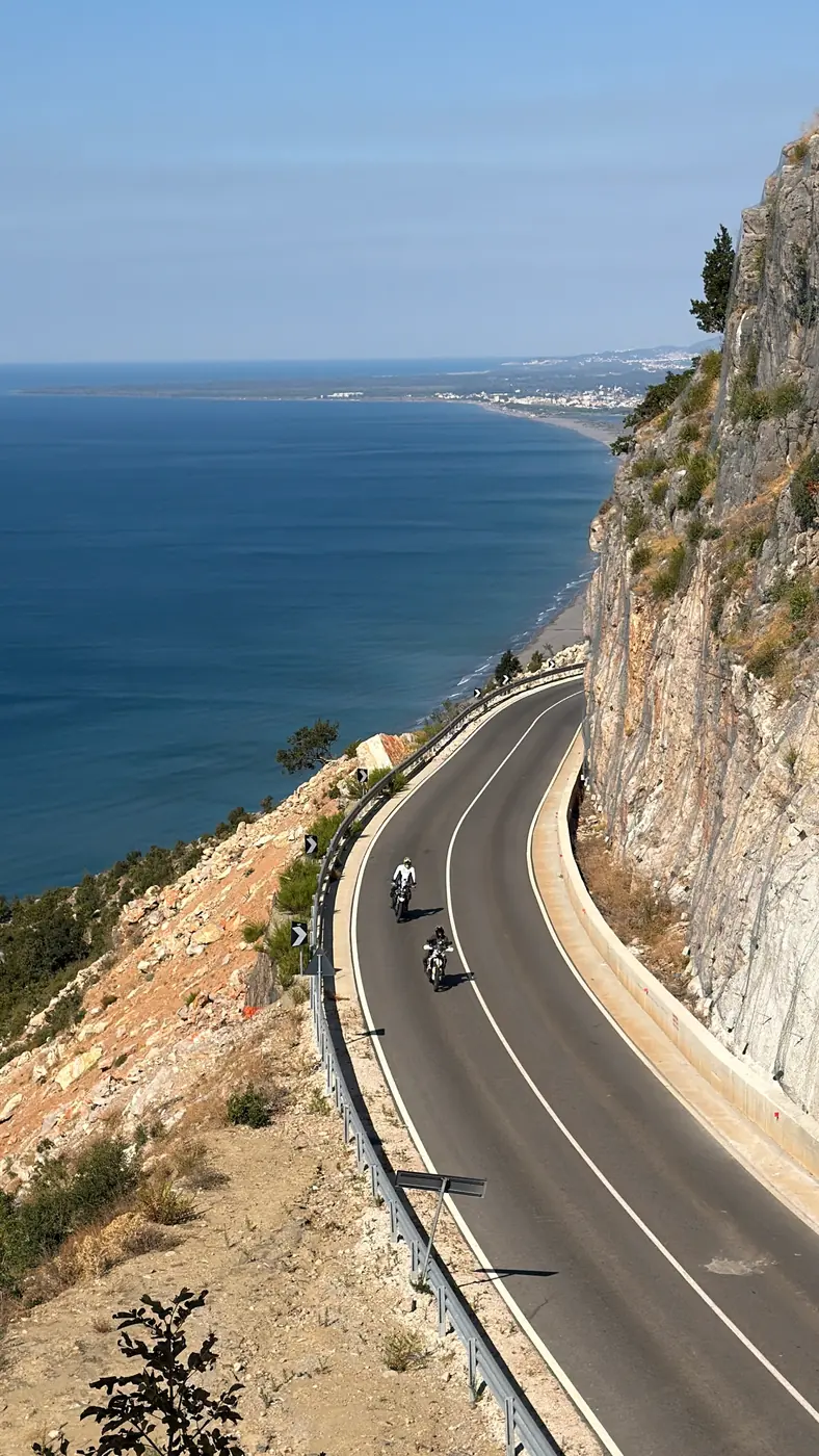 Two riders on a coast road cut into a cliff in southern Albania, the sea and a long beach far below