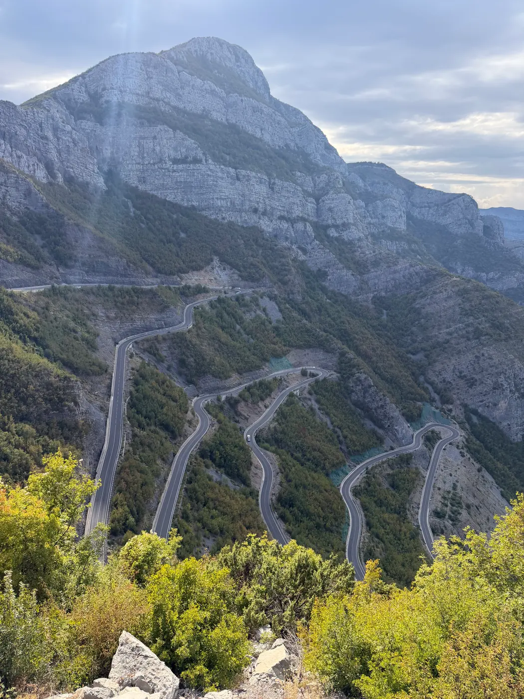 A road of tight hairpin switchbacks climbing a wooded gorge beneath a high limestone face in the Accursed Mountains, northern Albania
