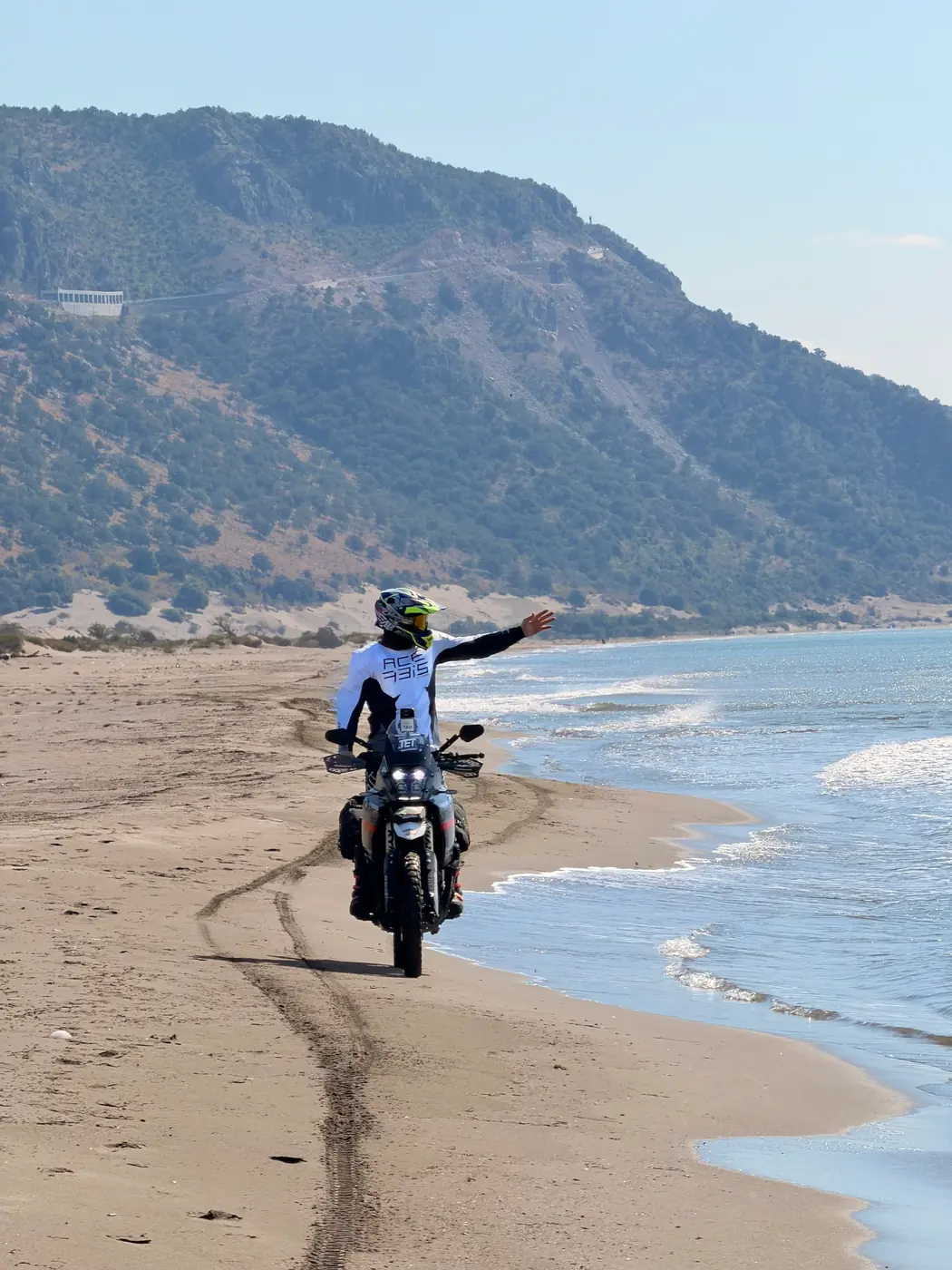 A rider on a loaded adventure bike riding along the sand at the water's edge in southern Albania, a green headland behind