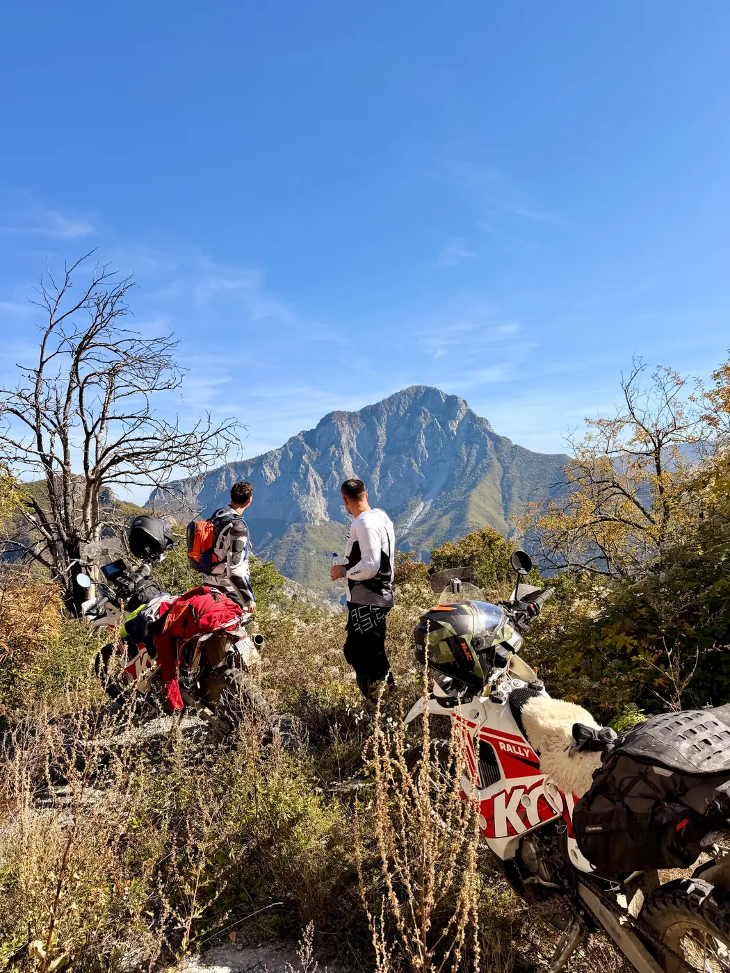 Two riders standing by their enduro bikes on a scrubby trail, facing a sharp limestone peak in the Accursed Mountains, northern Albania