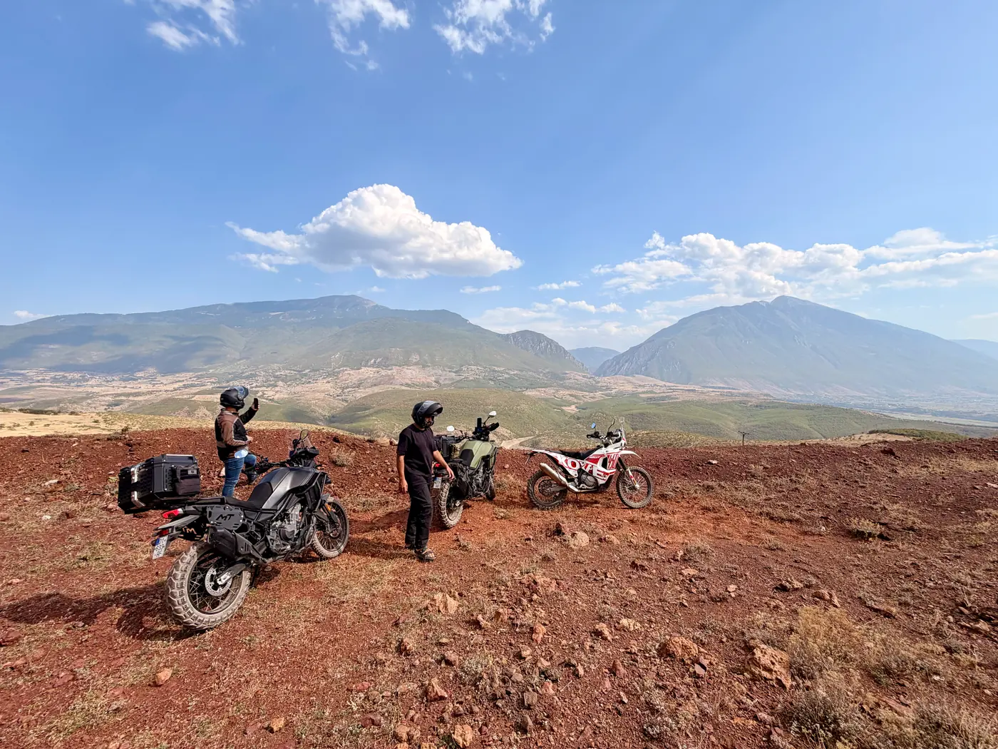 Three adventure bikes and two riders stopped on a red-earth ridge in southern Albania, with a wide valley and mountain ridges behind
